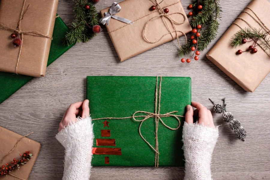 Hands in cozy white sleeves hold a green gift wrapped with twine on a table. Surrounding it are brown-paper packages, pine branches, and berries.