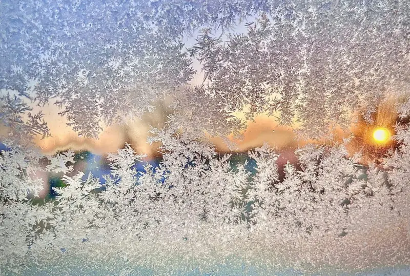 Frosted window with intricate ice patterns, revealing a soft, colorful sunrise in the background. The scene conveys a serene, chilly morning.