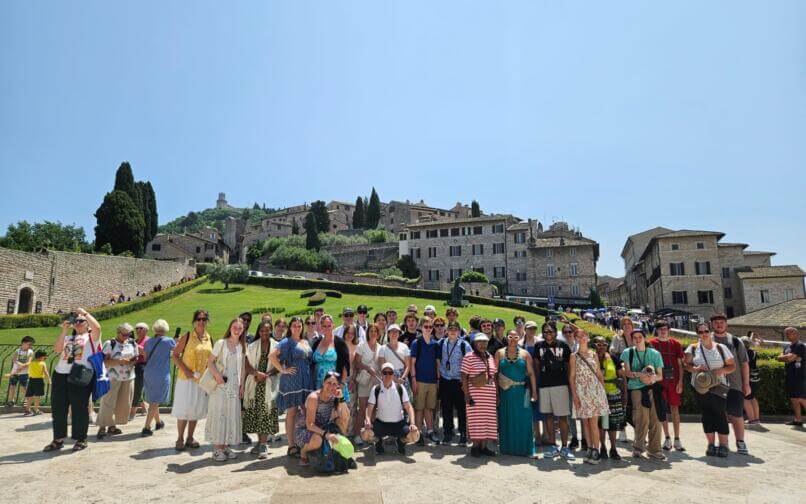 A large group of travelers posing together outdoors in front of a hillside town with stone buildings, green lawns, and tall cypress trees under a clear blue sky. The group stands closely together, smiling, with some people wearing hats, sunglasses, or backpacks.