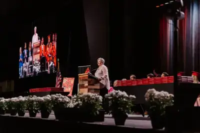 A speaker at a podium addresses a graduation ceremony. A large screen shows the same scene.