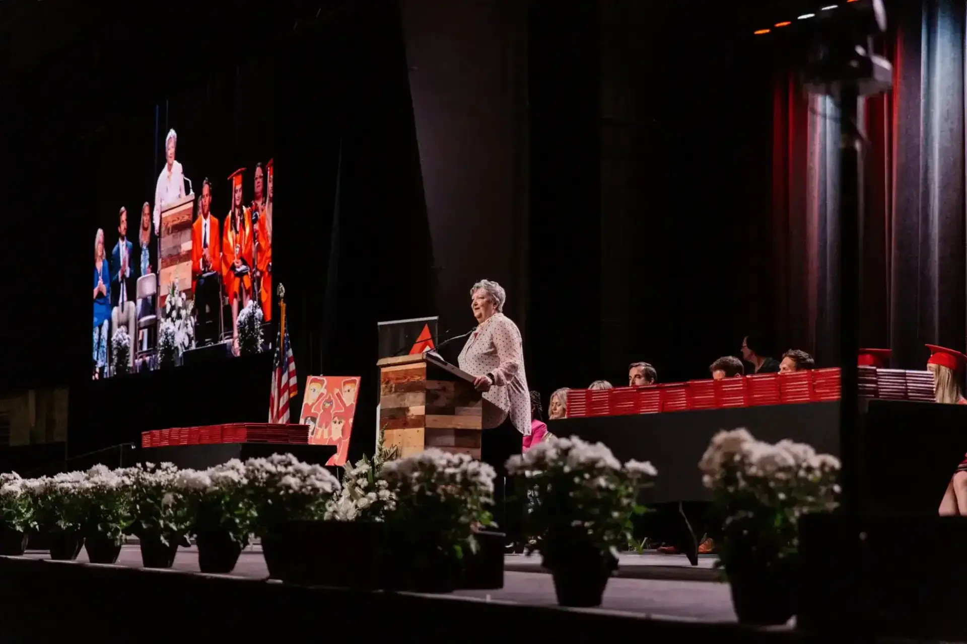 A speaker at a podium addresses a graduation ceremony. A large screen shows the same scene.