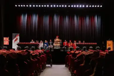 Graduation ceremony with students in red caps and gowns seated facing a stage. A speaker at the podium is flanked by faculty.