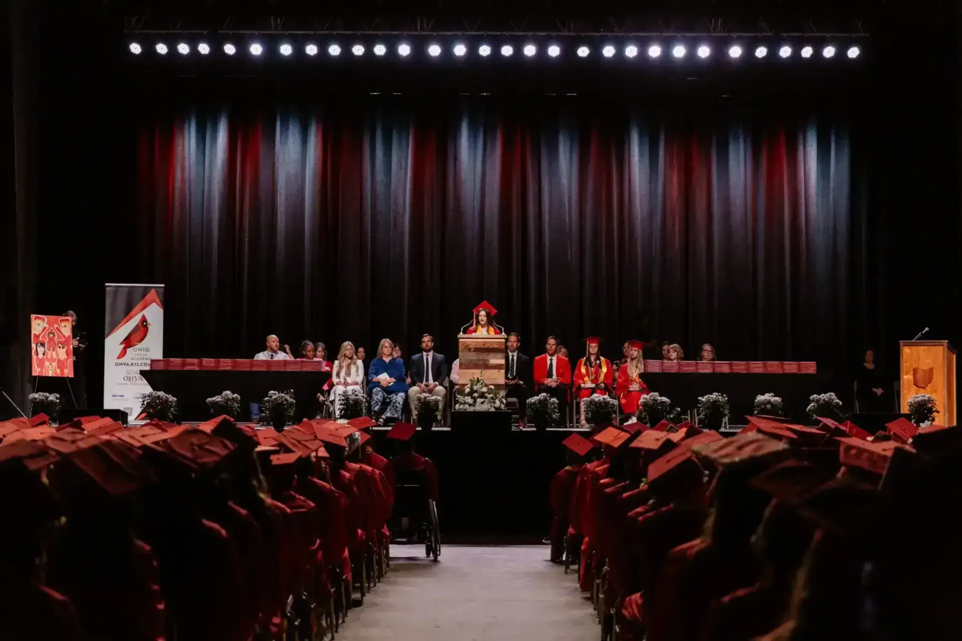 Graduation ceremony with students in red caps and gowns seated facing a stage. A speaker at the podium is flanked by faculty.