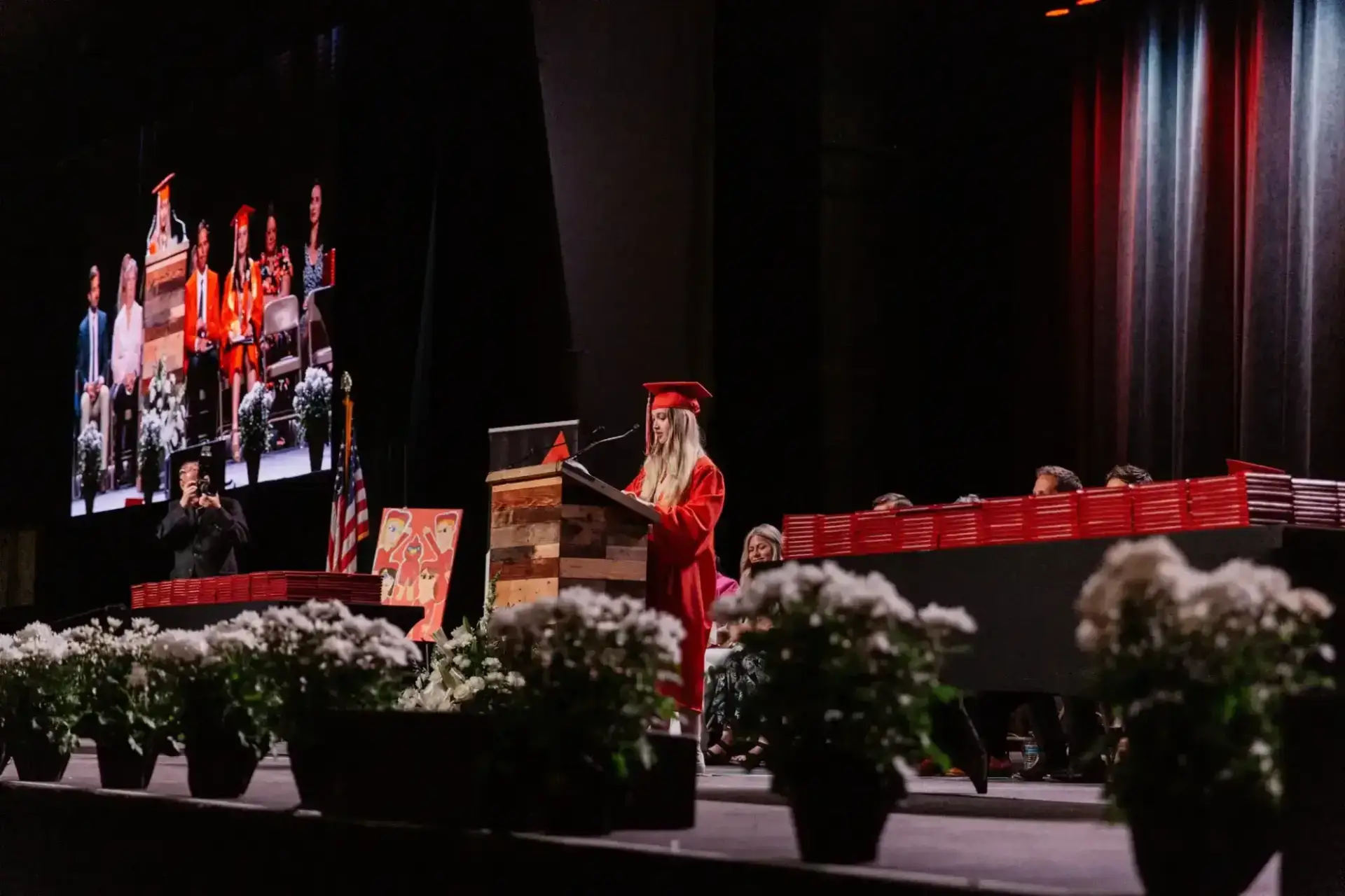 A student in a red cap and gown speaks at a wooden podium on a dimly lit graduation stage, with floral arrangements and a large screen backdrop.