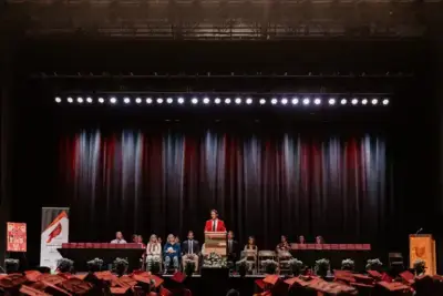 A speaker in a red robe stands at a podium on a stage, with an audience of graduates in red caps watching.