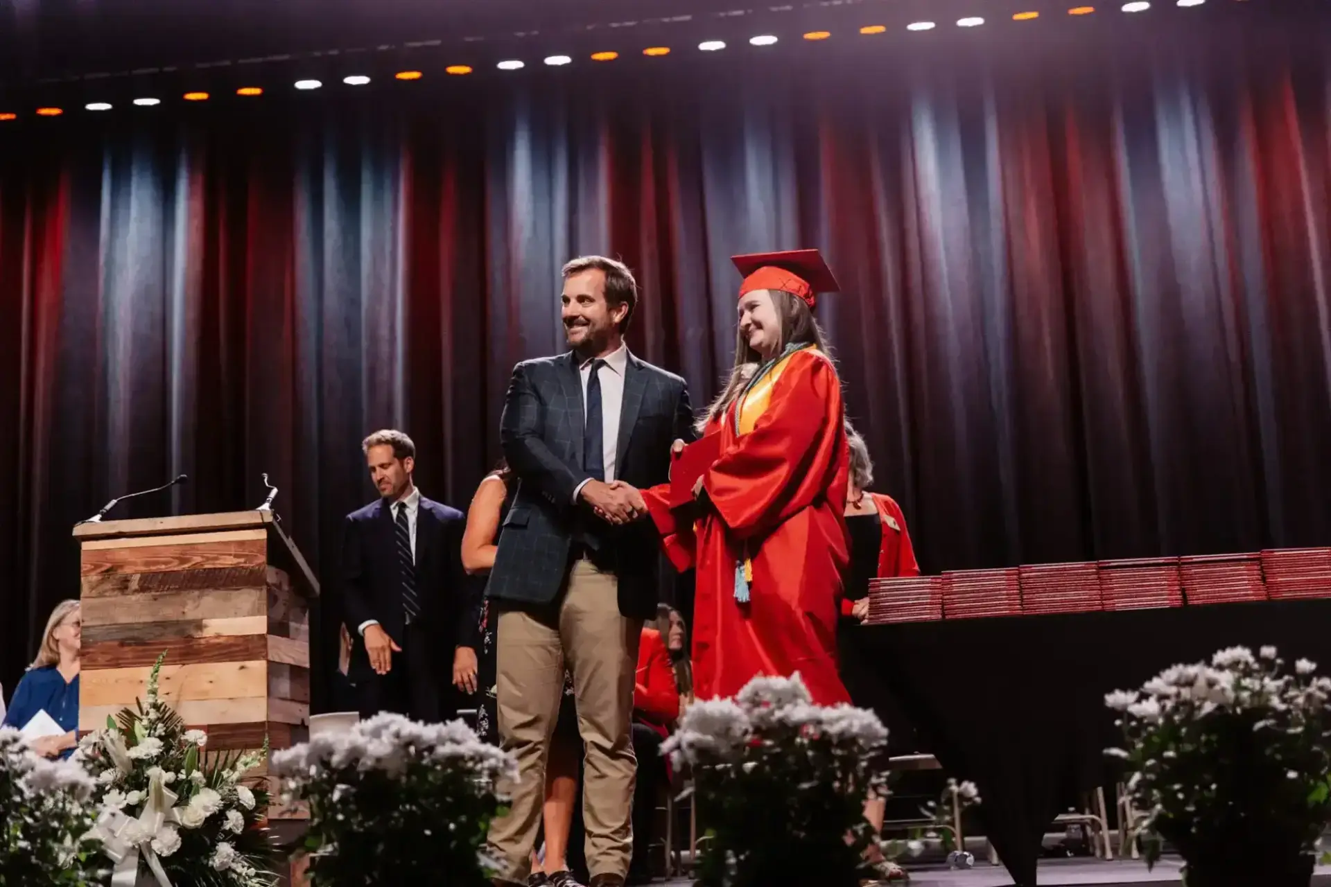 Graduate in red gown shakes hands with a smiling man at a graduation ceremony.