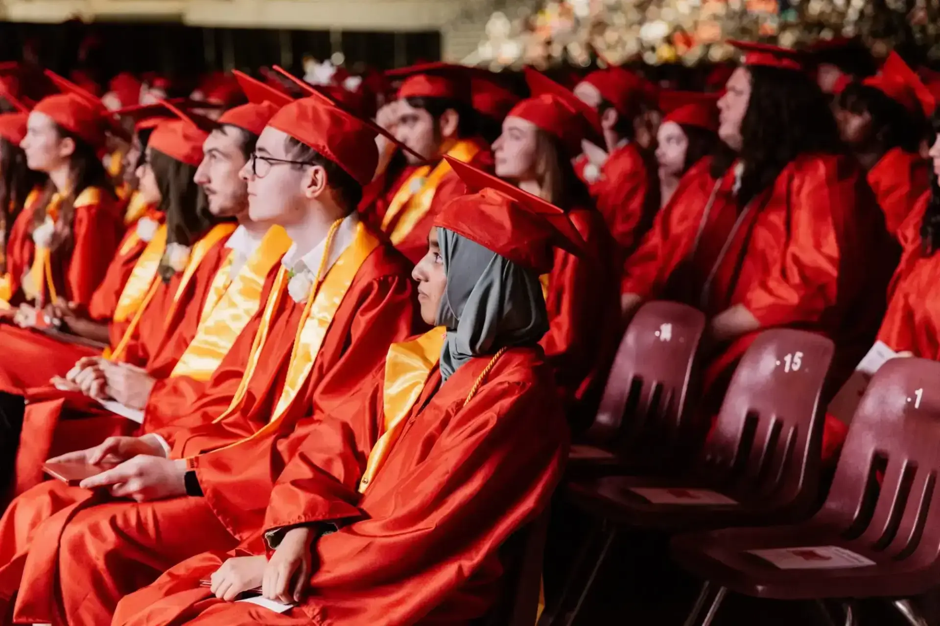 Graduates wearing red caps and gowns with yellow stoles sit in rows during a ceremony.