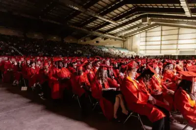 A large group of students in red caps and gowns sit in rows during a graduation ceremony inside a spacious auditorium filled with spectators.
