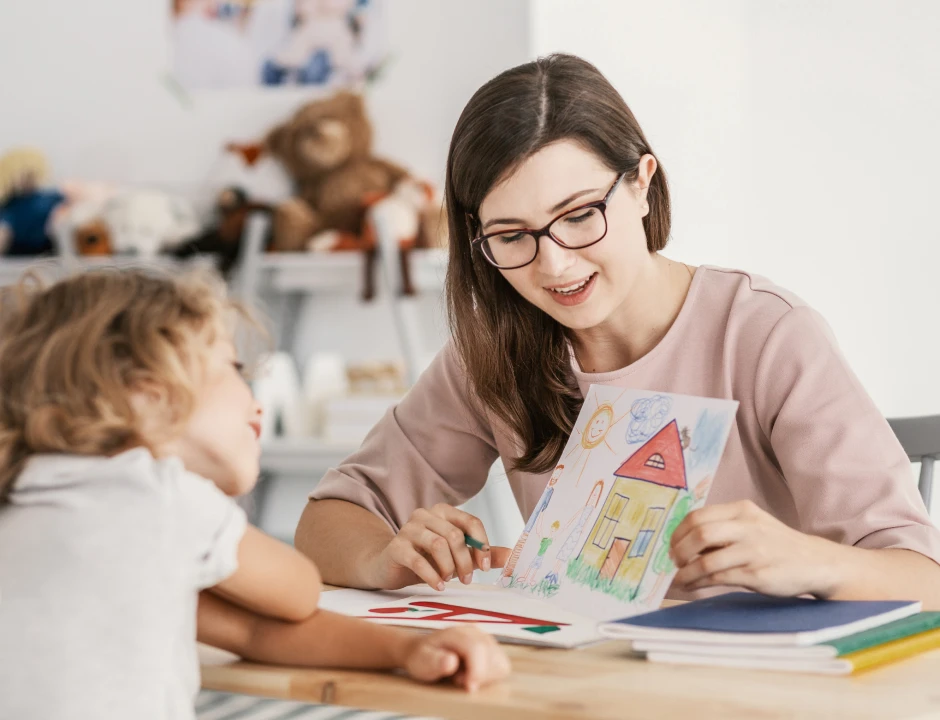 A smiling woman with glasses shows a colorful house drawing to a young child who watches intently. The scene is warm and nurturing, set in a playroom with toys.