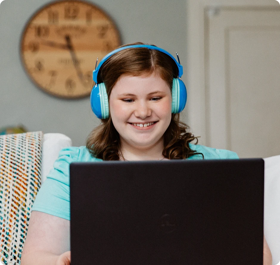 Smiling girl with blue headphones uses a laptop in a cozy room. A round wall clock and a patterned blanket are visible in the background.