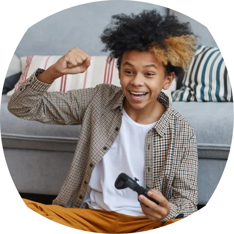 A young person with a checkered shirt and curly hair excitedly holds a game controller, fist raised, sitting on a couch with striped cushions.