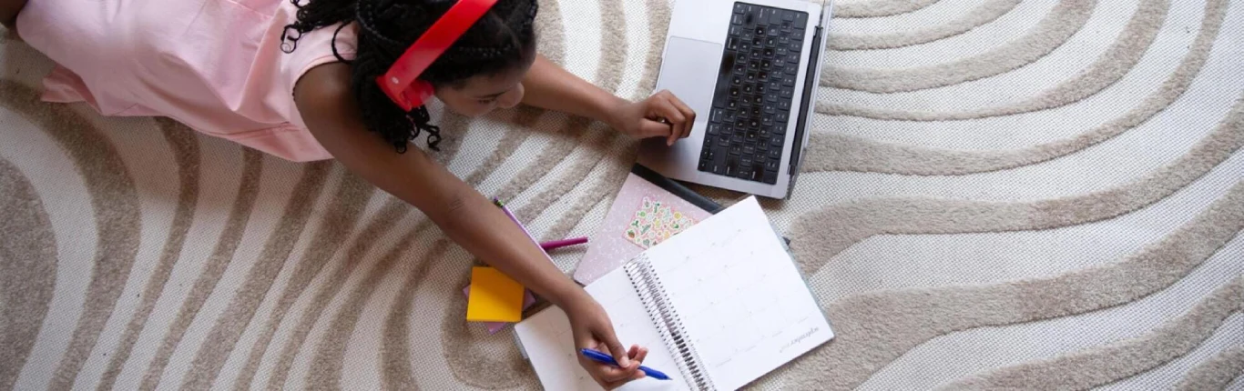 A child in a pink dress and red headphones lies on a patterned rug, using a laptop, writing in a planner, surrounded by colorful pens. She appears engaged.